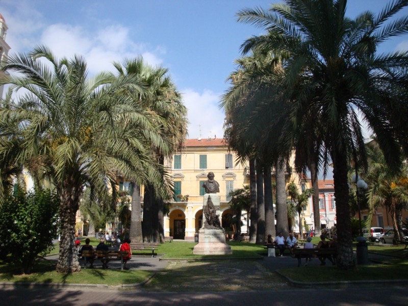 Giardino con palme a Diano Marina con il monumento ad Andrea Rossi e un palazzo giallo