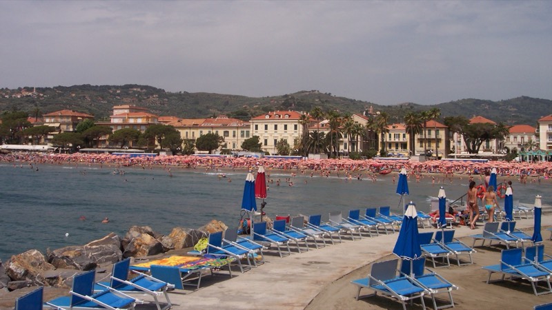 Spiaggia di Diano Marina d'estate con lettini blu, ombrelloni e bagnanti, lungomare e colline alle spalle
